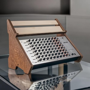Puede incluir: Un soporte de madera para mezclador de audio con una placa frontal plateada y numerosas perillas negras. El soporte está hecho de madera clara y tiene un diseño inclinado. El mezclador está sobre una mesa de vidrio.