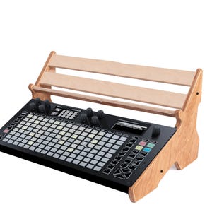 Könnte beinhalten: Schwarzes elektronisches Musikproduktionsinstrument mit einem Raster aus beleuchteten Tasten, Knöpfen und einem Bildschirm. Es ist auf einem hellen Holzständer mit Lattenrost montiert, der für den Studio- oder Live-Einsatz konzipiert ist.