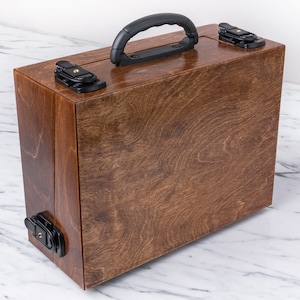 Peut inclure: Une bo&icirc;te de transport en bois avec une finition teint&eacute;e brune et une poign&eacute;e en plastique noir. La bo&icirc;te a deux loquets noirs de chaque c&ocirc;t&eacute;.