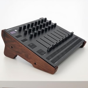 May include: A black Launch Control MIDI controller with multiple knobs, sliders, and buttons, mounted on a dark brown wooden stand. The controller features a small screen and is designed for music production.