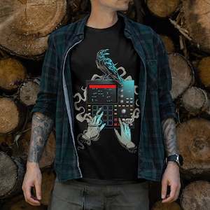 May include: Black t-shirt featuring a graphic of a blue crow perched on a black drum machine with two skeletal hands reaching towards the buttons. The drum machine has a red and blue display.
