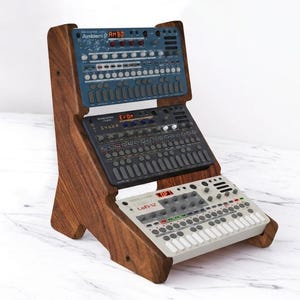 Pode incluir: Um suporte de madeira de três níveis que contém três sintetizadores de música eletrónicos. A unidade superior é azul, a do meio é cinzento-escura e a inferior é branca. Cada dispositivo possui numerosos botões circulares e ecrãs.