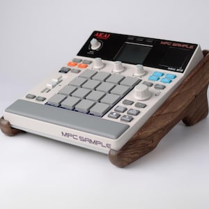 May include: A white and gray MPC Sample music production device with orange and blue buttons, and a black panel with the AKAI logo. The device is mounted on a wooden stand. The text "MPC SAMPLE" is visible on the device.