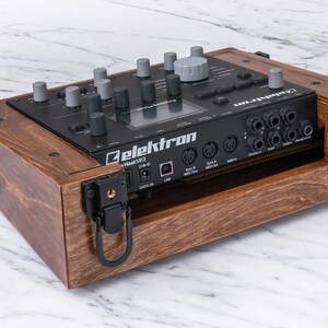 Peut inclure: Un bo&icirc;tier en bois avec des loquets et des poign&eacute;es noirs pour un synth&eacute;tiseur Elektron Heat MKII noir. Le bo&icirc;tier est en bois fonc&eacute; avec une finition naturelle.