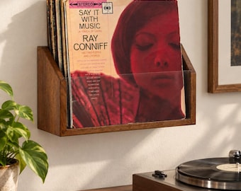 May include: A wall-mounted wooden record display shelf holding several vinyl albums. The front-most album cover features a red-toned portrait of a person and the text Say It With Music by Ray Conniff. The shelf has a clear acrylic front panel.