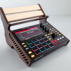 Puede incluir: Un centro de producción musical Akai MPC One negro con un soporte de madera. El dispositivo tiene una pantalla que muestra una interfaz de producción musical, con pads y botones coloridos. El soporte es de madera oscura y clara.