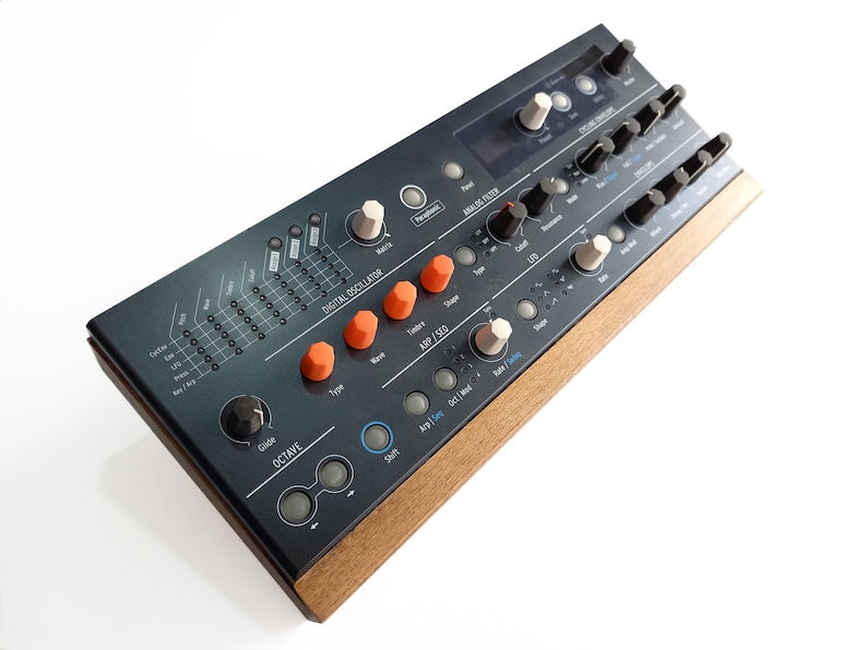 May include: A black and silver synthesizer with orange and white knobs. The synthesizer has a digital oscillator, an ARP/SEQ section, and an analog filter. The synthesizer is made of metal and has a wooden side panel.