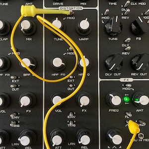 May include: A close-up of a black synthesizer with knobs, buttons, and a yellow patch cable connecting the different modules. The synthesizer has labels such as "Drive", "Distortion", "Mod", "Tune", "Warp", "HPF", "BPF", "FX", "Env", "Trig", "Att", "Rel", "Rec", "Play", "SD", "HH", "CV", "Freq", "Amt", "Dly", "Rev", "CLK Mod", "FB", "MAD", "OPCH", "MIDI Volume", and "PWR".
