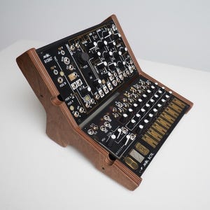 Puede incluir: Un sintetizador modular con marco de madera. El panel negro presenta numerosas perillas, interruptores y conectores. El marco de madera es de color marrón cálido, y el sintetizador está inclinado para facilitar el acceso.