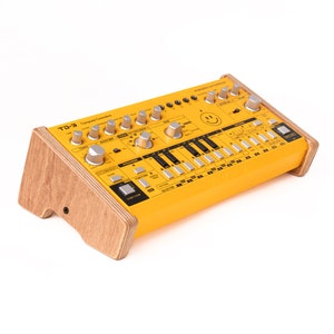 May include: A yellow analogue synthesizer with a wooden stand. The synthesizer has a smiley face on the front and the text "Analog/Sync Synthesizer" above the smiley face. The text "TD-3" is on the left side of the synthesizer.