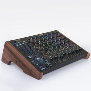 Puede incluir: Un mezclador de audio negro con numerosas perillas, botones y controles deslizantes. El mezclador tiene un marco de madera y está sobre un fondo blanco. La parte superior del mezclador tiene una cuadrícula de luces multicolores.