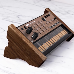 May include: A KORG volca keys synthesizer in a wooden stand. The synthesizer is copper colored with black keys and knobs. The stand is made of dark brown wood.