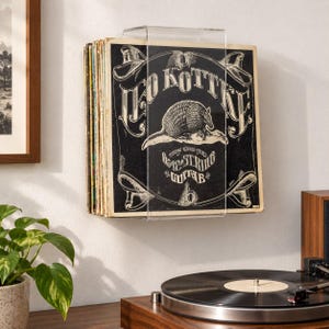 May include: A clear acrylic wall-mounted vinyl record display shelf holding a collection of records. The front-most record features a black and white cover with an armadillo illustration and the text Leo Kottke 6- and 12-String Guitar.