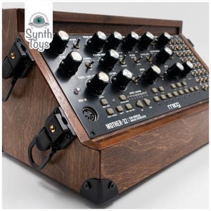 May include: A dark brown wooden case with black metal accents houses a Moog Mother-32 semi-modular analog synthesizer. The synthesizer has a black faceplate with knobs, buttons, and a patch bay.