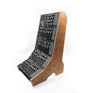 Op de afbeelding: Een modulair synthesizersysteem met een houten frame. De zwarte bedieningspanelen zijn schuin geplaatst voor gemakkelijke toegang, met talrijke knoppen en schakelaars. Het lichtbruine houten frame heeft zichtbare houtnerf.