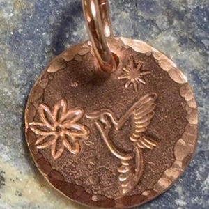 May include: A round copper pendant featuring a hammered edge and a stamped design of a hummingbird, a flower, and a small star. The pendant includes a circular jump ring at the top for attachment, showcasing a warm, metallic reddish-brown finish.