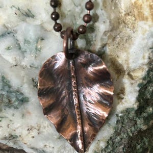 May include: A copper leaf pendant necklace. The pendant is a detailed, textured leaf shape with a central vein. The necklace has a ball chain and a copper finish. The leaf pendant is approximately 2 inches long.