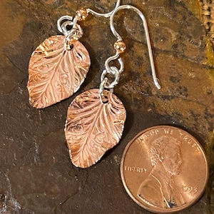 May include: A pair of copper-colored leaf-shaped earrings with intricate embossed patterns, suspended from silver-toned metal hooks. A standard United States one-cent coin is positioned next to the earrings to provide a clear sense of scale.