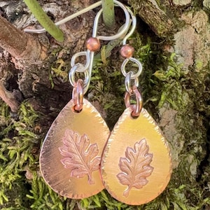 May include: A pair of teardrop-shaped copper earrings featuring a stamped oak leaf design. The earrings hang from silver-toned metal hooks and include small copper beads at the top of the jump rings, showcasing a metallic finish.