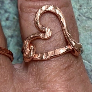 May include: A hammered copper ring in the shape of a heart. The ring has a textured surface and is worn on a finger. The heart shape is formed by the ring's design, with the top of the heart curving inward.