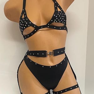 May include: Black two-piece lingerie set with rhinestone embellishments. The halter top and high-waisted bottoms are connected by a belt and leg straps. The set is displayed on a mannequin.