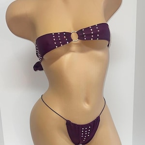 Puede incluir: Un top y una parte inferior de bikini morado con pedrería. La parte superior es sin tirantes y tiene un anillo en el centro. La parte inferior es de estilo tanga.