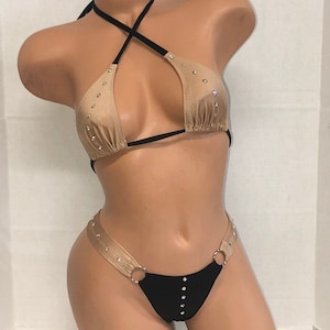 May include: A two-piece swimsuit with a black and nude color scheme. The top is a halter style with a criss-cross strap and the bottom is a cheeky style with a single ring on each side. Both pieces are adorned with rhinestones.