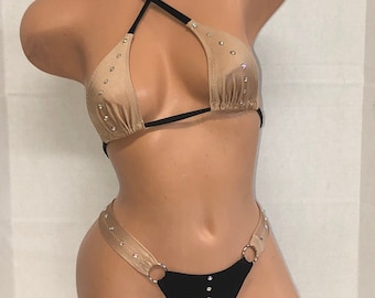 Puede incluir: Un traje de baño de dos piezas en color negro y nude. La parte superior es de estilo halter con una correa cruzada y la parte inferior es de estilo atrevido con un solo anillo a cada lado. Ambas piezas están adornadas con pedrería.
