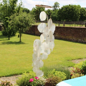 Peut inclure: Un carillon à vent en coquillages blancs avec un anneau en bois au sommet. Le carillon est suspendu dans un jardin avec de l'herbe verte et des arbres en arrière-plan.