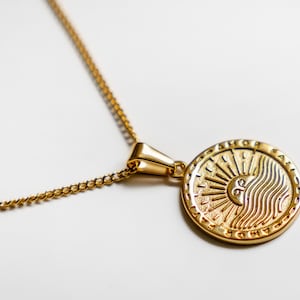 May include: A gold chain necklace with a round pendant featuring a sun and waves design. The pendant has a textured surface and is surrounded by a raised border.