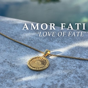 Może przedstawiać: Złoty naszyjnik z okrągłym wisiorem przedstawiającym słońce i fale. Wisiorek jest ozdobiony napisem "Amor Fati" i "Love of Fate".