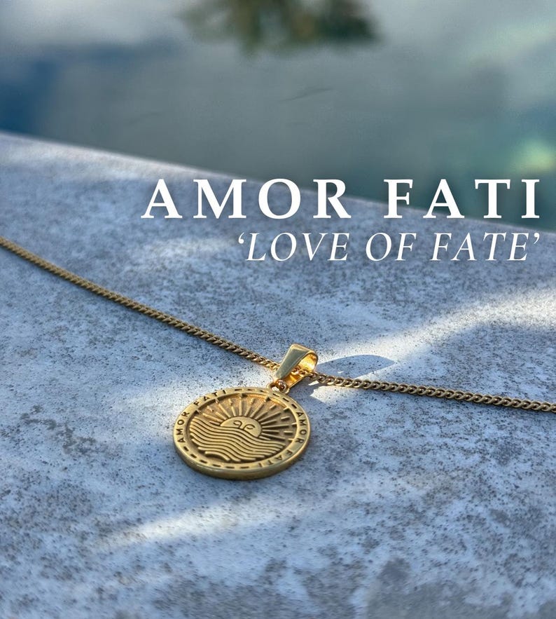 Może przedstawiać: Złoty naszyjnik z okrągłym wisiorem przedstawiającym słońce i fale. Wisiorek jest ozdobiony napisem "Amor Fati" i "Love of Fate".