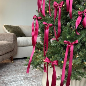 May include: A Christmas tree decorated with burgundy satin ribbon bows. The tree is in a burgundy planter with a ribbed design.