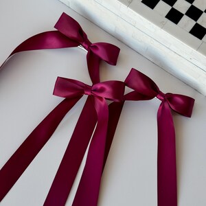 May include: Three burgundy satin ribbon hair bows with long tails. The bows are tied with a silver clip.