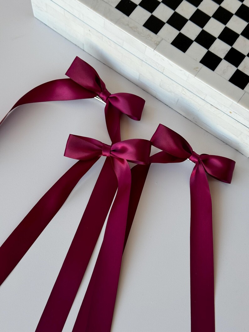 May include: Three burgundy satin ribbon hair bows with long tails. The bows are tied with a silver clip.