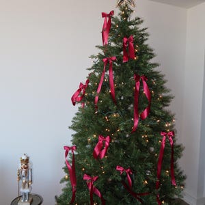 May include: A decorated Christmas tree with a gold star on top and red satin ribbon bows. The tree is lit with white lights and is in a room with white walls.