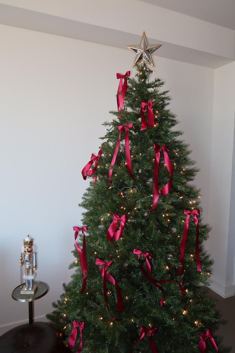 May include: A decorated Christmas tree with a gold star on top and red satin ribbon bows. The tree is lit with white lights and is in a room with white walls.