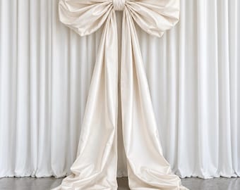 May include: A large, decorative bow made of smooth, cream-colored fabric. The bow features two voluminous, gathered loops at the top and two long, flowing fabric tails that extend downward, pooling slightly at the base of the display.