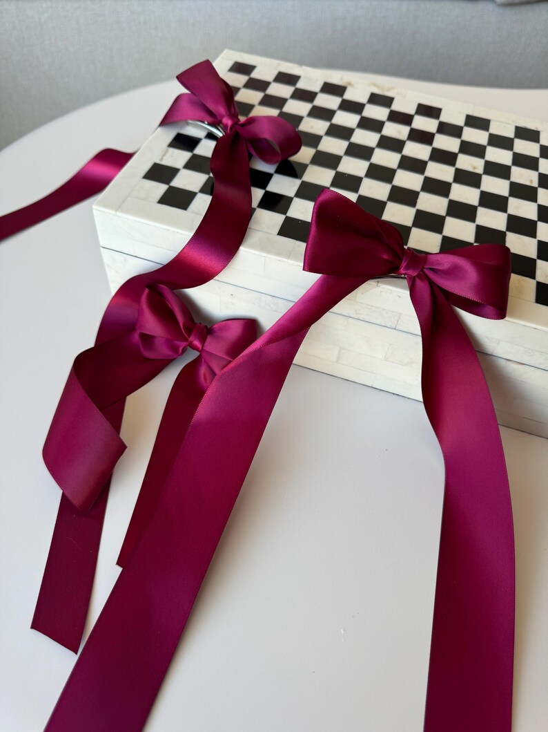 May include: Three burgundy satin ribbon bows tied on a black and white checkered box. The box is made of white marble with black squares.