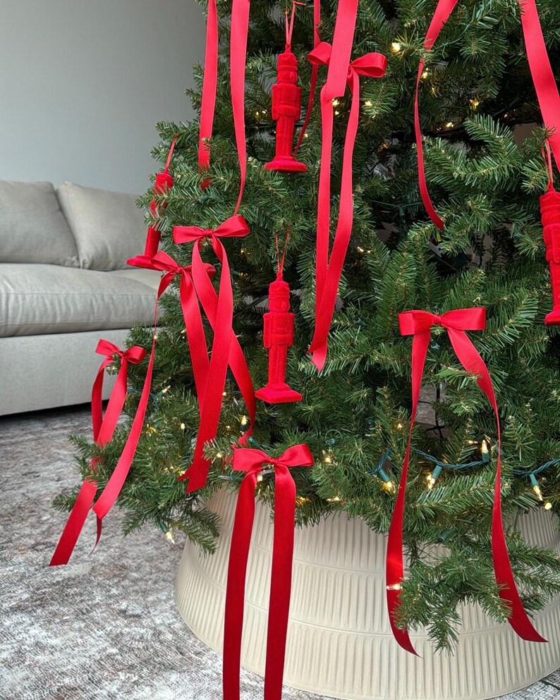 May include: A Christmas tree decorated with red velvet nutcracker ornaments and red satin ribbon bows. The tree is in a white planter with a textured design.