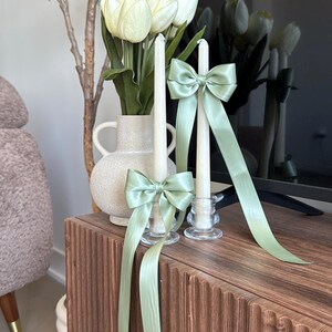 May include: Two white candles with sage green satin bows tied around them. The candles are in glass candle holders and are sitting on a wooden table.