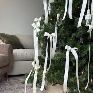 May include: A Christmas tree decorated with white satin ribbon bows. The tree is in a white planter and is lit with white Christmas lights.