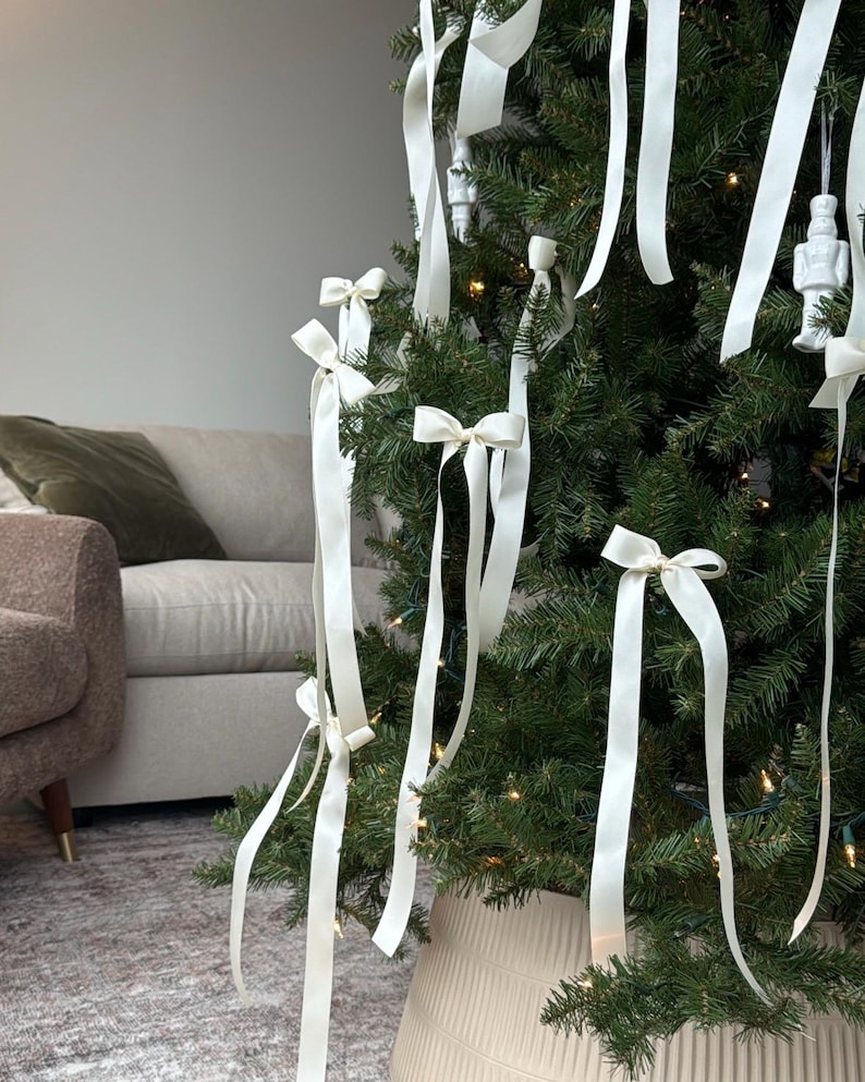 May include: A Christmas tree decorated with white satin ribbon bows. The tree is in a white planter and is lit with white Christmas lights.