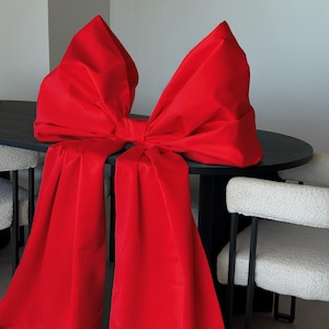 May include: A large, vibrant red bow made of fabric, draped over a black table. The bow is the focal point, with its voluminous shape and flowing tails. The background includes white chairs and a neutral wall.