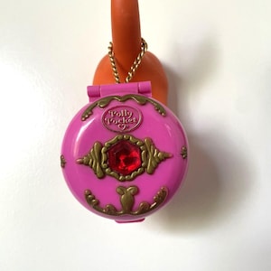 Może przedstawiać: Różowy kompakt Polly Pocket ze złotymi akcentami i czerwonym klejnotem. Okrągły kompakt ma pokrywę na zawiasach i złoty łańcuszek. Tekst "Polly Pocket" jest widoczny na pokrywie.