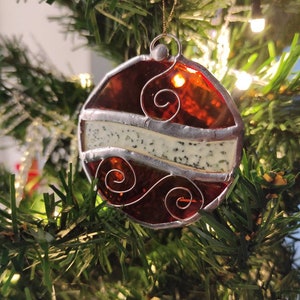 May include: A stained glass Christmas ornament with a red and white design. The ornament has a silver frame and a white background with a red stripe and swirls.