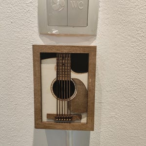 May include: A framed wooden art piece depicting a laser-cut acoustic guitar. The guitar has light-coloured wood with dark brown details, set against a white background. The frame is a warm brown, mounted on a white wall. Above it is a light switch.