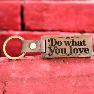 Puede incluir: Llavero de madera con una correa de cuero marrón y un anillo de latón. La etiqueta de madera tiene el texto "Do what you love" grabado.