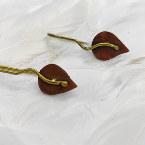 以下が含まれることがあります： 茶色の金属仕上げと金色のワイヤーアクセントが付いた、小さな葉の形をしたイヤリングのペア。イヤリングは白い背景の上にあります。