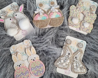 May include: Five pairs of beaded earrings displayed on white cards with the word "Boujee" printed on them. The earrings are shaped like a bunny, a basket, and a flower. The bunny earrings feature pastel pink, purple, and green beads. The basket earrings are made with gold, pink, green, and white beads. The flower earrings are adorned with white, pink, and gold beads.